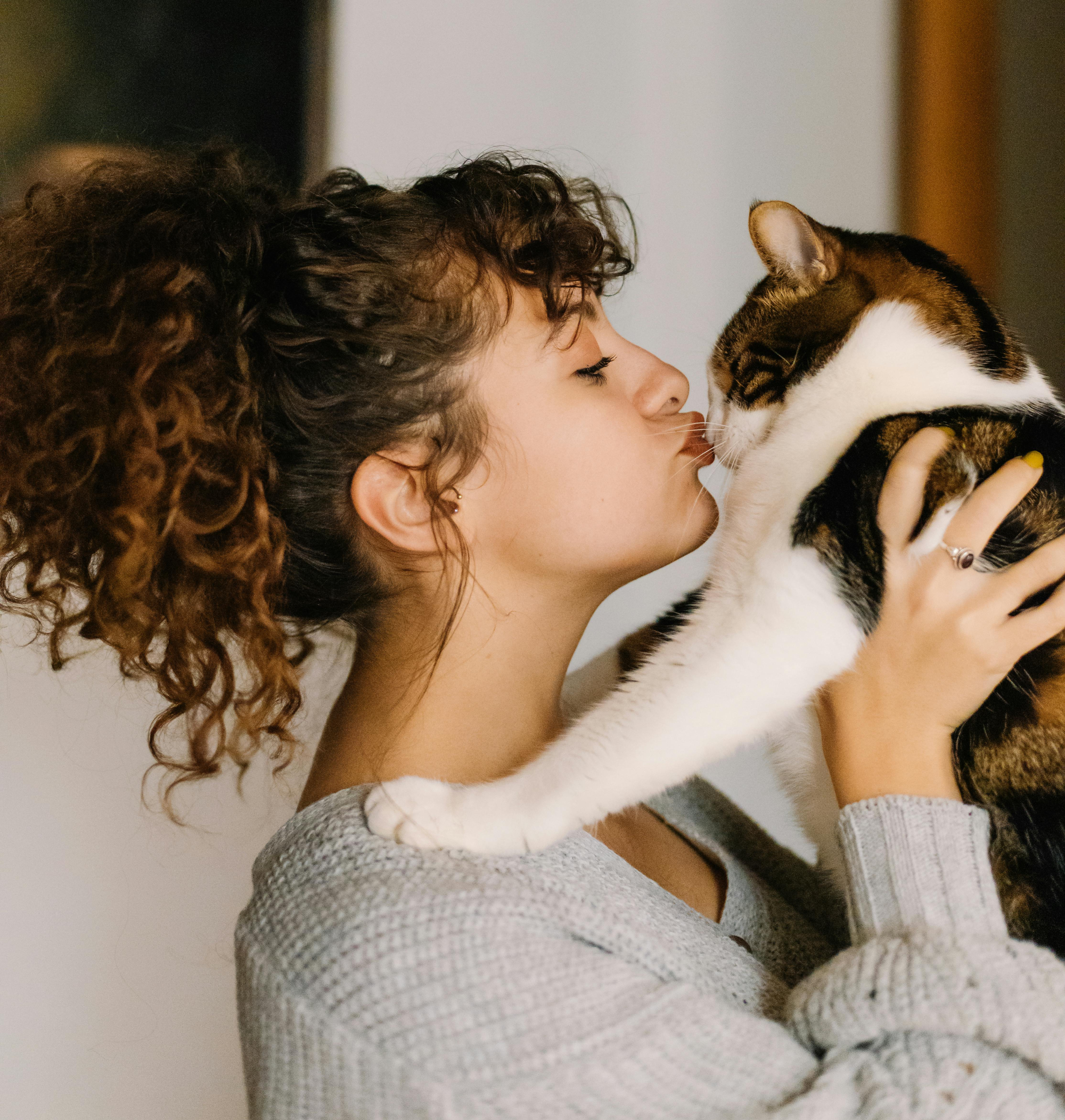 Junge Frau küsst ihre Katze im Arm – liebevoller Moment zwischen Mensch und Tier in gemütlicher Wohnumgebung