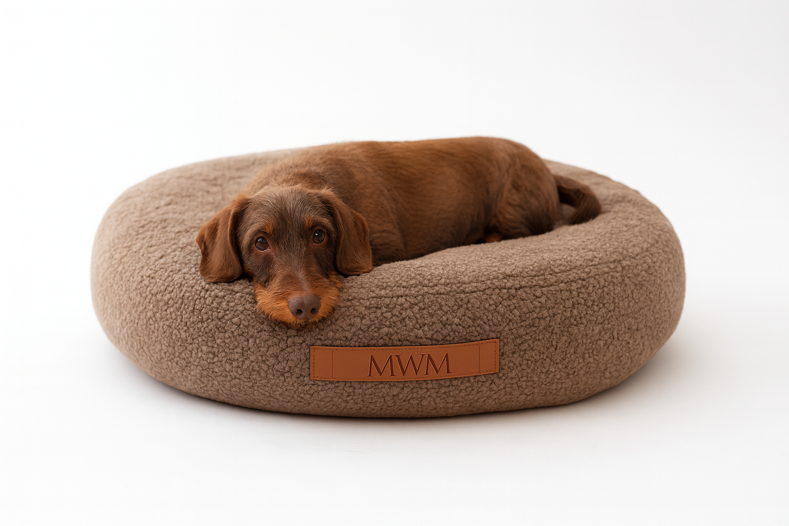 Dackel liegt entspannt in einem runden, waschbaren Hundebett aus braunem Bouclé-Stoff mit MVM-Logo – ideal für kleine bis mittelgroße Hunde.